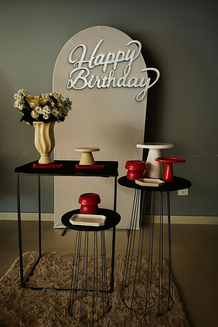 Painel bege com letreiro Happy Birthday, mesas pretas altas e peças em vermelho e cru