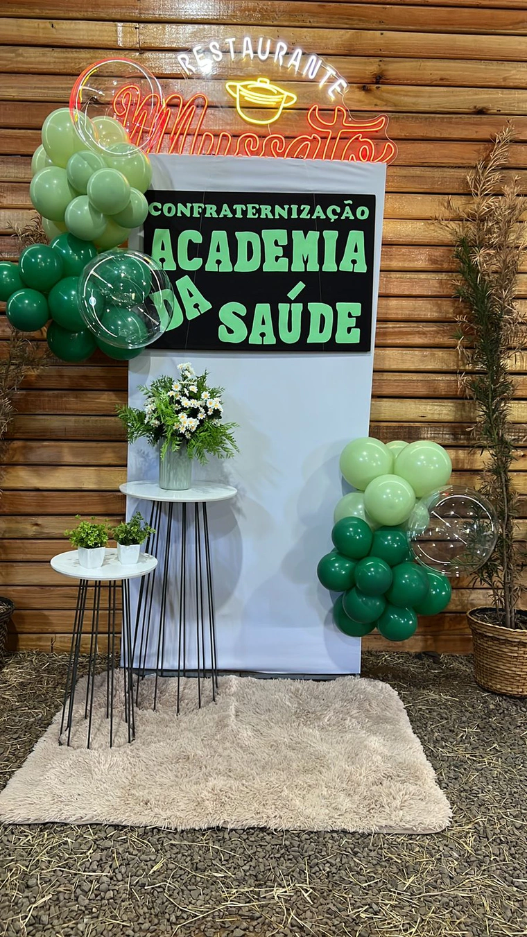 Painel branco com faixa de confraternização, balões verdes e mesas altas com flores